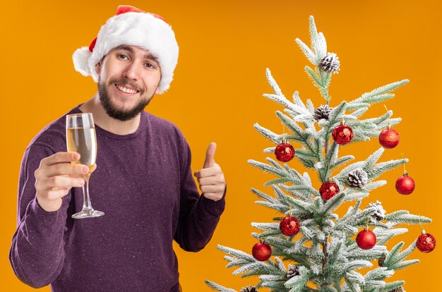 Joven en suéter morado y gorro de Papá Noel sosteniendo una copa de champán junto al árbol de Navidad sobre fondo naranja