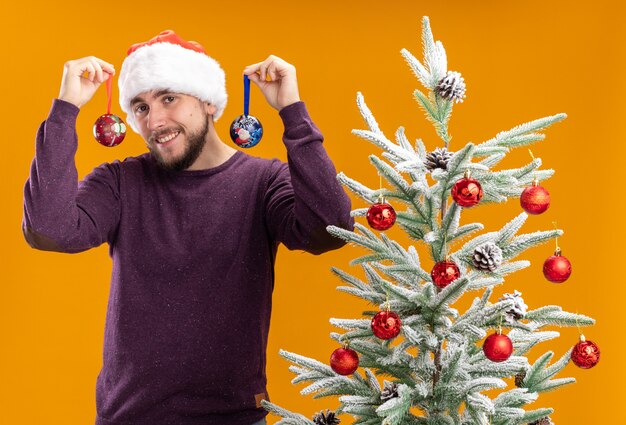 Joven en suéter morado y gorro de Papá Noel sosteniendo juguetes mirando a la cámara con cara feliz sonriendo de pie junto al árbol de Navidad sobre fondo naranja