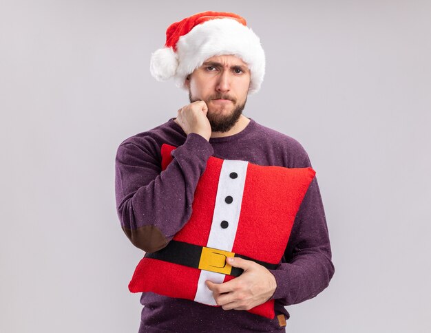 Joven en suéter púrpura y gorro de Papá Noel sosteniendo una almohada divertida mirando a la cámara disgustado de pie sobre fondo blanco.