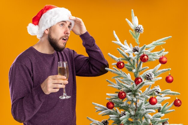 Joven en suéter púrpura y gorro de Papá Noel sosteniendo una copa de champán de pie junto al árbol de Navidad mirando hacia arriba desconcertado sobre la pared naranja