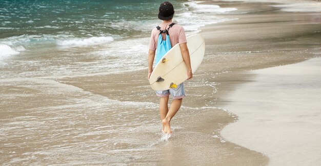 Joven surfista descalzo con mochila caminando por la playa, con bodyboard blanco debajo del brazo, regresando a casa después de un viaje intenso