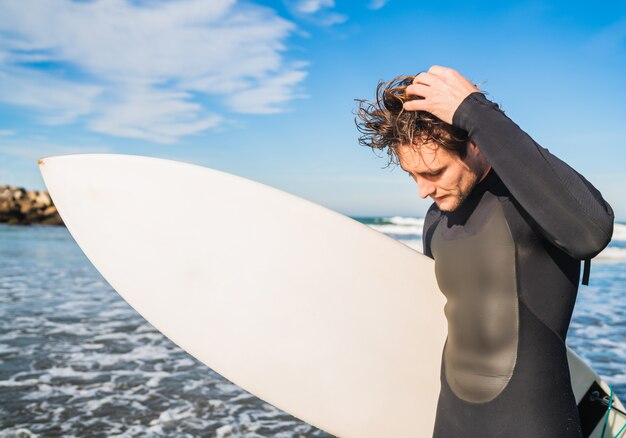 Joven surfista de pie en el océano con su tabla de surf en un traje de surf negro. Concepto de deporte y deporte acuático.