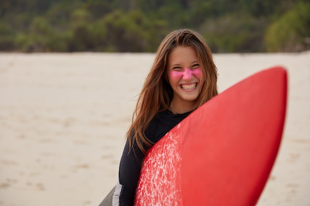 Joven surfista profesional alegre sonríe ampliamente, lleva una tabla de surf encerada roja, tiene zinc en la cara, feliz después de golpear una gran ola del océano, modelos contra la pared de arena, expresa positividad