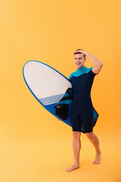 Joven surfista sonriente caminando con tabla de surf y mirando a otro lado