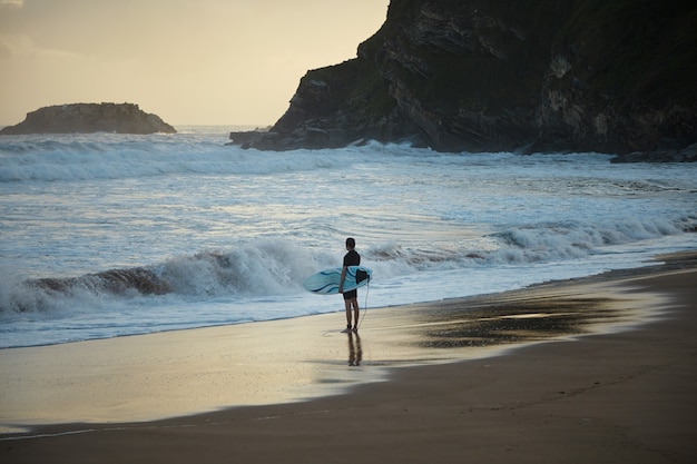 Joven surfista en traje de neopreno corto con funboard en mano permanece solo en la playa de surf oculta al amanecer listo para ir al océano
