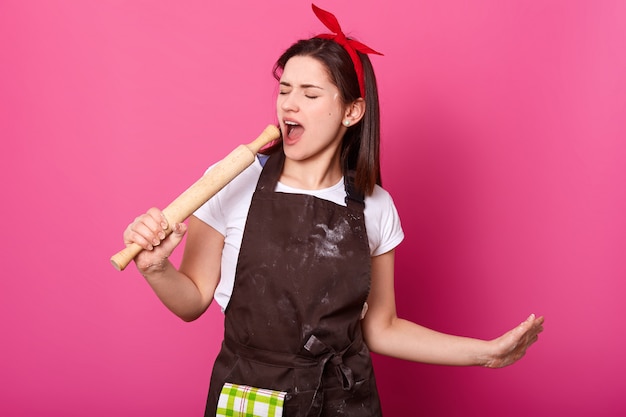 Joven talentosa sostiene un rodillo en su mano fingió su micrófono, abre la boca mientras canta. Linda dama enérgica usa delantal marrón, camiseta blanca y diadema roja. Concepto de cocina