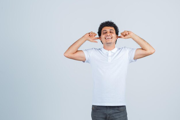 Joven tapando los oídos con los dedos índices, cerrando los ojos con camiseta blanca y jeans y luciendo feliz, vista frontal.