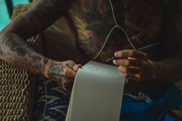 un joven tatuado con auriculares escucha música y dibuja un cuaderno.