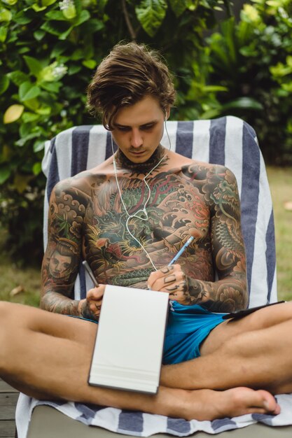 un joven tatuado con auriculares escucha música y dibuja un cuaderno.