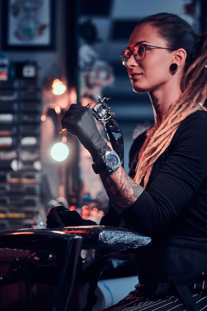 Joven tatuadora atractiva con guantes negros está afinando su máquina de tatuajes antes de la sesión de tatuajes.