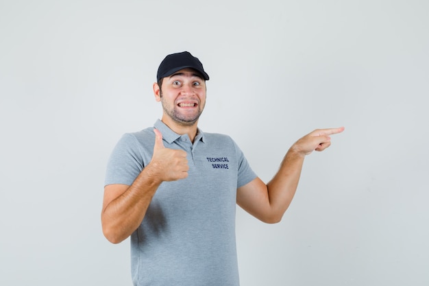 Joven técnico apuntando a un lado, mostrando el pulgar hacia arriba en uniforme gris y mirando contento.