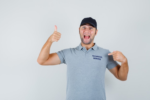 Joven técnico apuntando a su camiseta, mostrando el pulgar hacia arriba en uniforme y luciendo feliz.