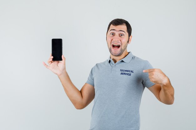 Joven técnico apuntando a este teléfono con los dedos índices en uniforme gris y mirando sorprendido, vista frontal.