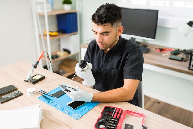 Joven técnico masculino que usa aire comprimido para limpiar dentro de un teléfono inteligente sucio en el taller de reparación. Hombre latino con guantes limpiando un celular