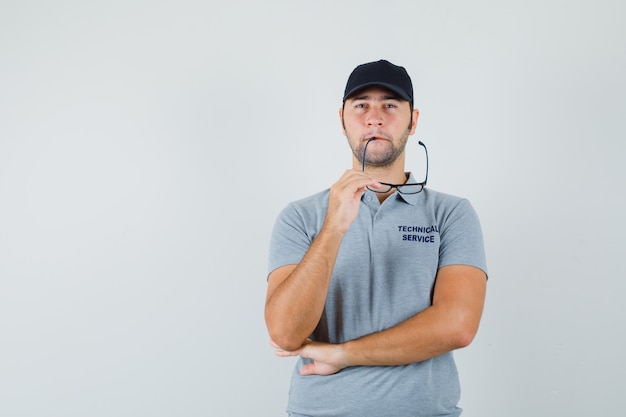 Joven técnico mordiendo vasos en uniforme gris y mirando vacilante.