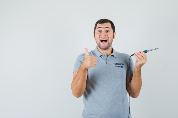 Joven técnico sosteniendo un destornillador y mostrando el pulgar hacia arriba en uniforme gris y mirando satisfecho.