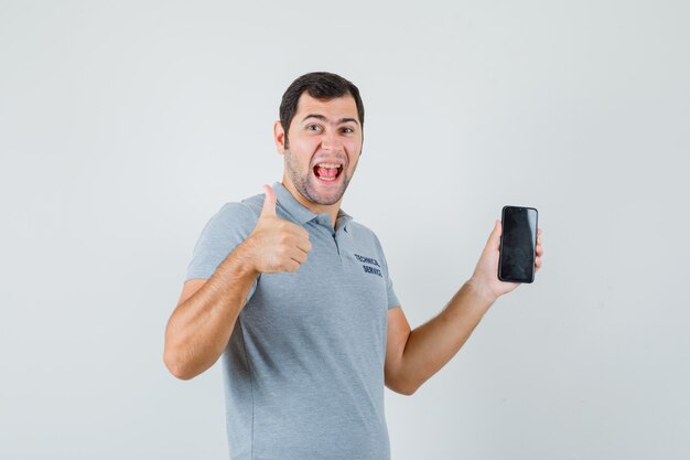 Joven técnico sosteniendo el teléfono móvil, mostrando el pulgar hacia arriba en uniforme gris y luciendo feliz, vista frontal.