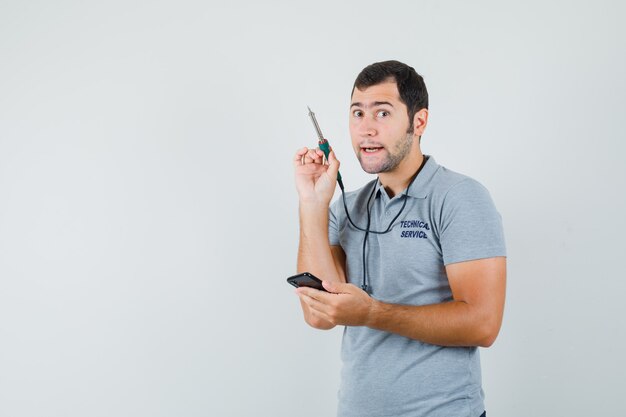 Joven técnico tratando de abrir su teléfono inteligente usando un taladro en uniforme gris y mirando enfocado.