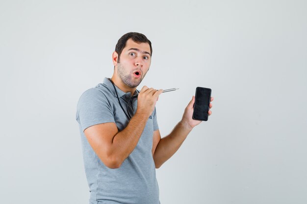 Joven técnico tratando de abrir su teléfono inteligente usando un taladro en uniforme gris y mirando sorprendido, vista frontal.