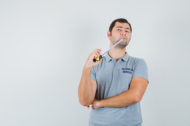 Joven técnico en uniforme gris mordiendo cinta métrica y de pie en pose de pensamiento y mirando pensativo.