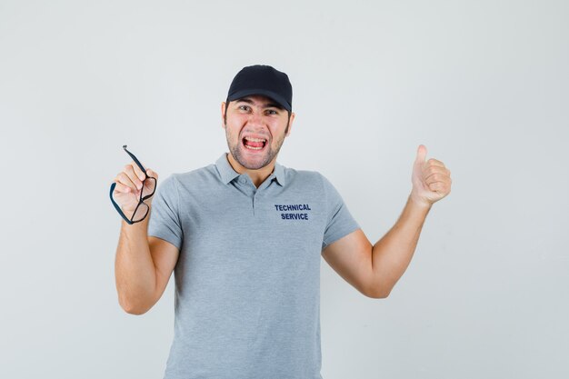 Joven técnico en uniforme gris mostrando el pulgar hacia arriba, sosteniendo vasos y mirando feliz.