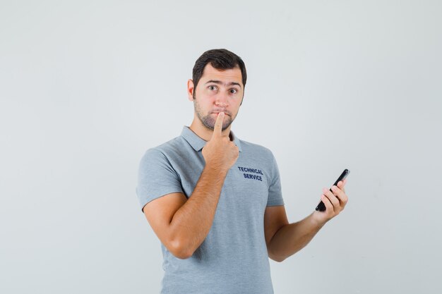 Joven técnico en uniforme gris de pie en pose de pensamiento mientras sostiene el teléfono y mira pensativo.