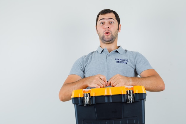 Foto gratuita joven técnico en uniforme gris sosteniendo la caja de herramientas con ambas manos y mirando sorprendido.