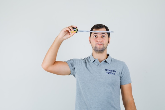 Joven técnico en uniforme gris sosteniendo cinta métrica contra su cabeza y mirando serio.