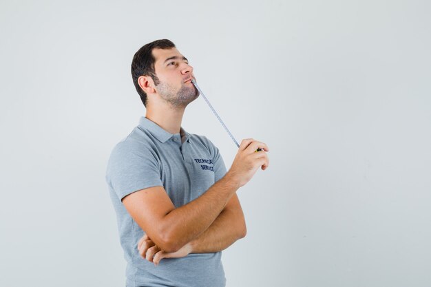 Joven técnico en uniforme gris sosteniendo cinta métrica en una mano y de pie en pose de pensamiento y mirando pensativo.