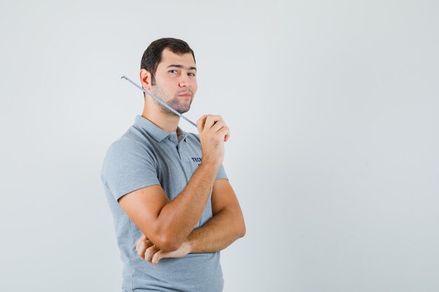 Joven técnico en uniforme gris sosteniendo cinta métrica y de pie en pose de pensamiento y mirando pensativo.