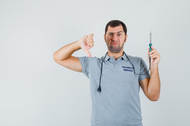 Joven técnico en uniforme gris sosteniendo el taladro en una mano mientras muestra el pulgar hacia abajo y parece disgustado.