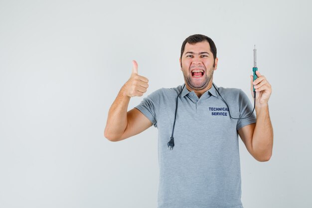 Joven técnico en uniforme gris sosteniendo el taladro en una mano mientras muestra el pulgar hacia arriba y se ríe y parece sorprendido.