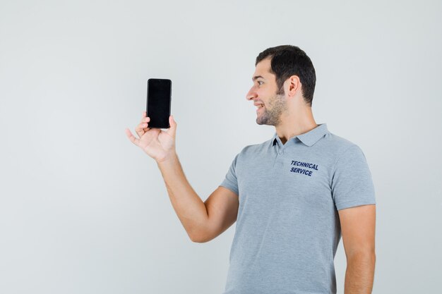 Joven técnico en uniforme gris sosteniendo el teléfono inteligente en la mano y sonriendo mientras lo mira y luce optimista, vista frontal.