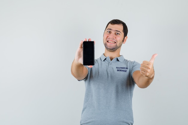 Joven técnico en uniforme gris sosteniendo el teléfono móvil, mostrando el pulgar hacia arriba y mirando alegre, vista frontal.