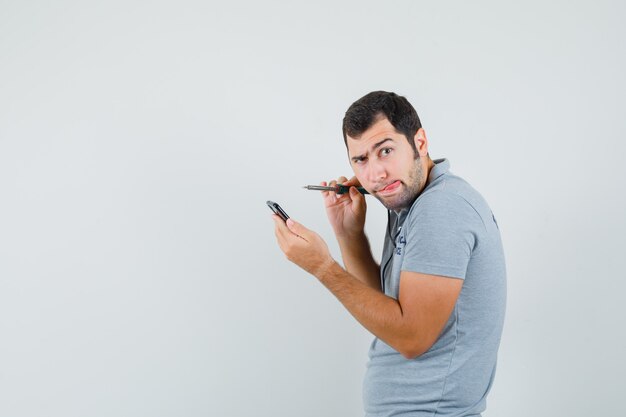 Joven técnico en uniforme gris tratando de abrir su teléfono inteligente usando un taladro y sacando la lengua y mirando enfocado.