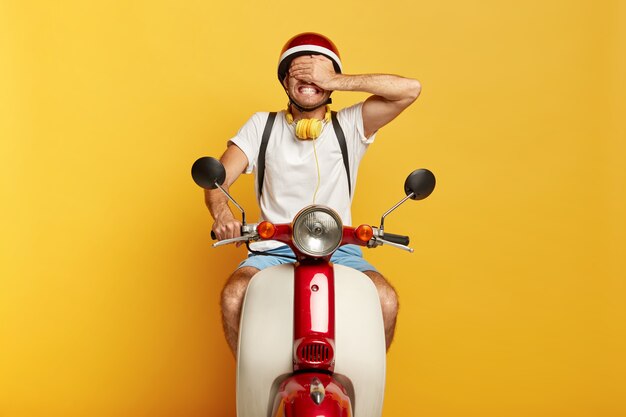 El joven tiene transporte propio, monta un scooter, se cubre los ojos con la palma, vestido con ropa casual, aislado sobre fondo amarillo