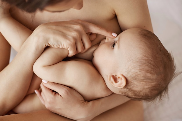 Joven tierna madre desnuda amamantando abrazando a su bebé recién nacido sentado en la cama por la mañana.