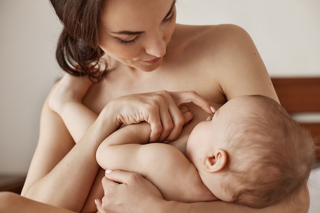 Joven tierna madre desnuda amamantando abrazando a su bebé recién nacido sentado en la cama por la mañana