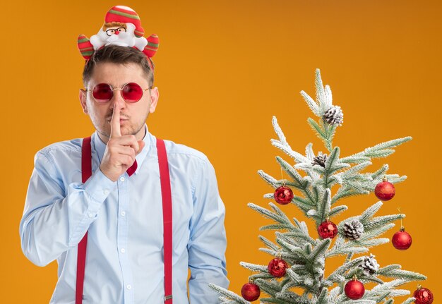 Joven con tirantes pajarita en el borde con santa y gafas rojas de pie junto al árbol de navidad haciendo gesto de silencio con el dedo en los labios sobre la pared naranja