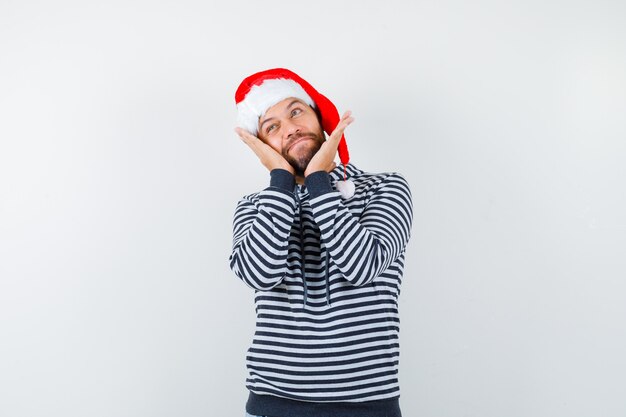 Joven tomados de la mano en la cabeza, mirando hacia arriba con capucha, gorro de Papá Noel y mirando curioso,