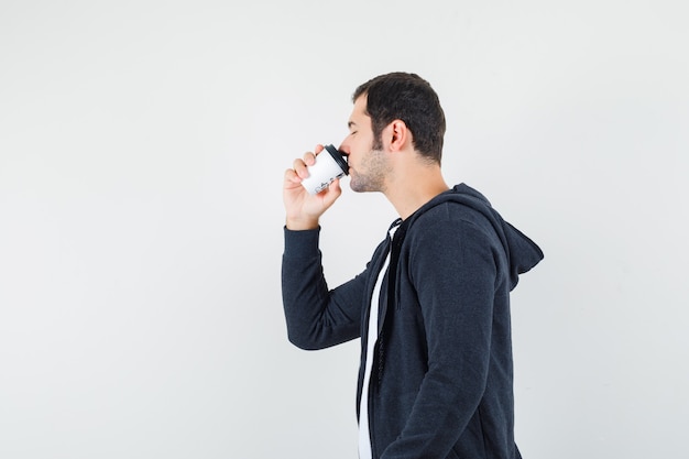 Joven tomando café en camiseta blanca y sudadera con capucha negra con cremallera frontal y mirando tranquilo, vista frontal.