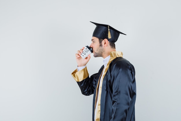 Joven tomando café en uniforme de posgrado y mirando pensativo.