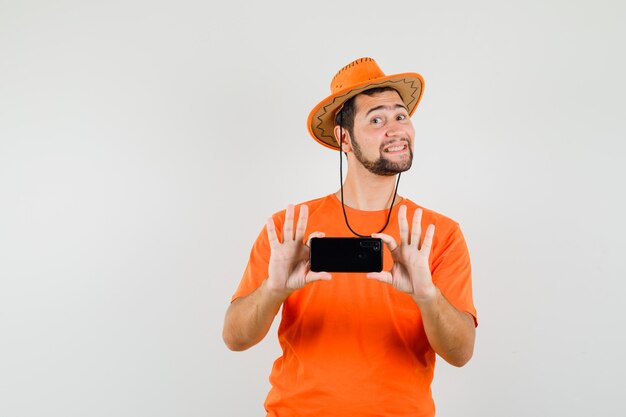 Joven tomando fotos en el teléfono móvil en camiseta naranja, sombrero y mirando alegre, vista frontal.