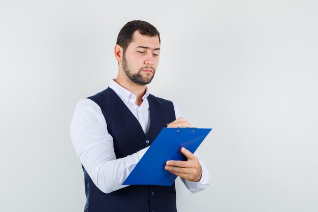 Joven tomando notas en el portapapeles en camisa y chaleco y mirando ocupado