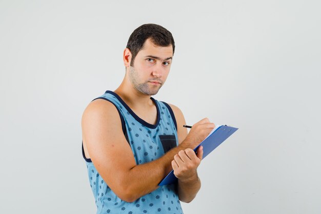 Joven tomando notas en el portapapeles en camiseta azul y mirando pensativo, vista frontal.