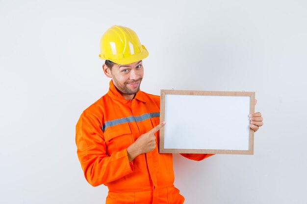 Joven trabajador apuntando al marco en blanco en uniforme, casco y mirando feliz.