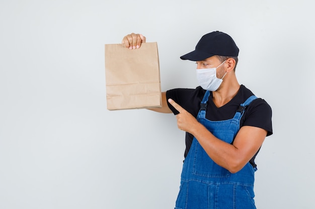 Joven trabajador apuntando a la bolsa de papel en uniforme, máscara, vista frontal.