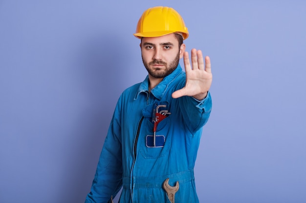 Joven trabajador barbudo guapo con casco amarillo y uniforme haciendo gesto de parada con su mano negando situación