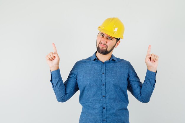 Joven trabajador en camisa, casco apuntando con el dedo hacia arriba y mirando insatisfecho