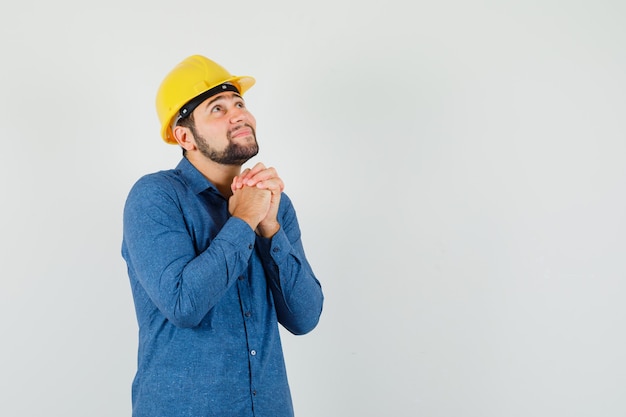 Foto gratuita joven trabajador en camisa, casco juntando las manos en gesto de oración y mirando esperanzado